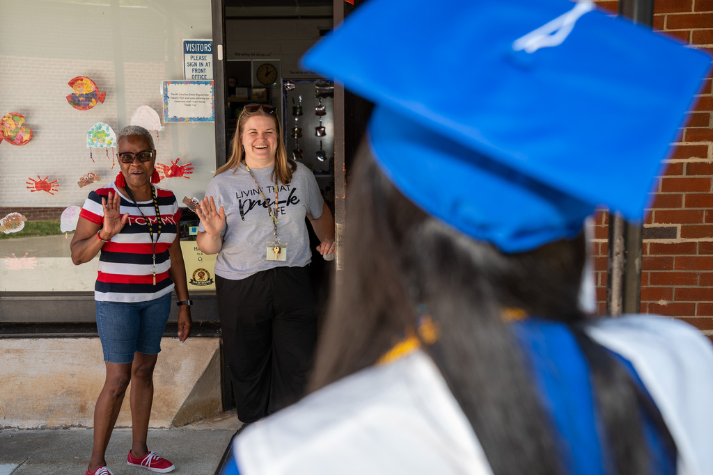Over-the-shoulder view of a graduate in a blue cap looking toward a school doorway. Two women stand at the entrance waving and smiling warmly. One woman wears a gray shirt that reads "Livin' That Pre-K Life."