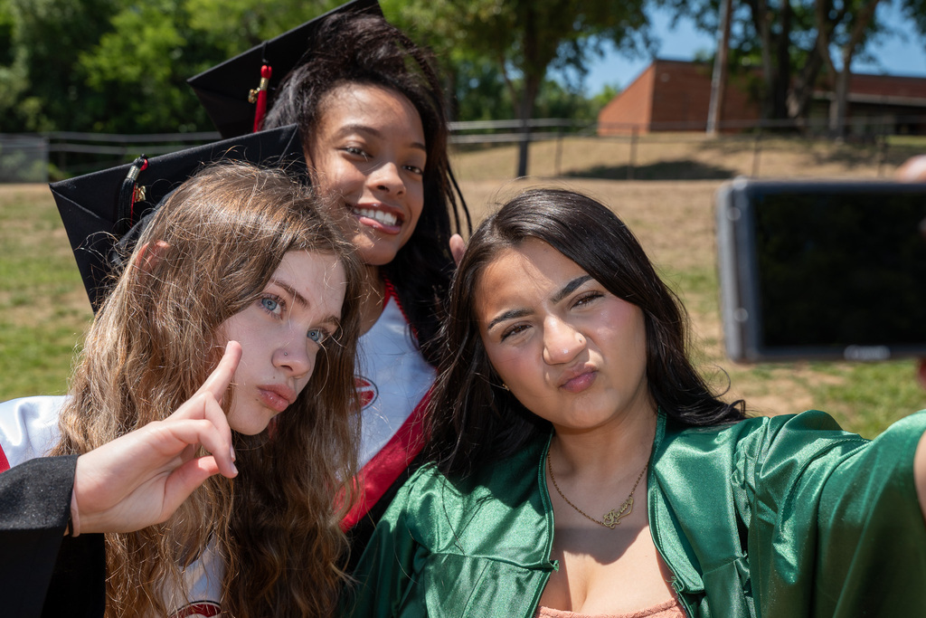Close-up selfie perspective of three female graduates posing outdoors. The girl on the right in a green gown holds the camera, making a pouty face. The girl in the center smiles brightly, and the girl on the left makes a peace sign toward her face.