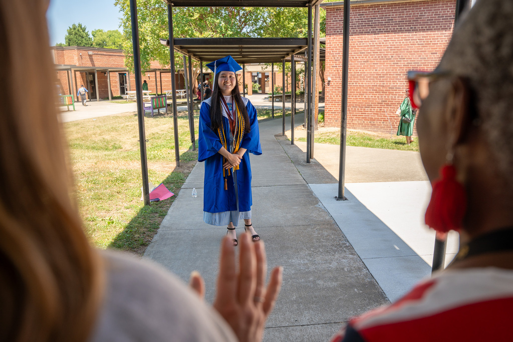A female graduate in a blue gown and cap stands smiling under an outdoor covered walkway at a school. The photo is framed from behind the shoulders of two women in the foreground who are waving to her.