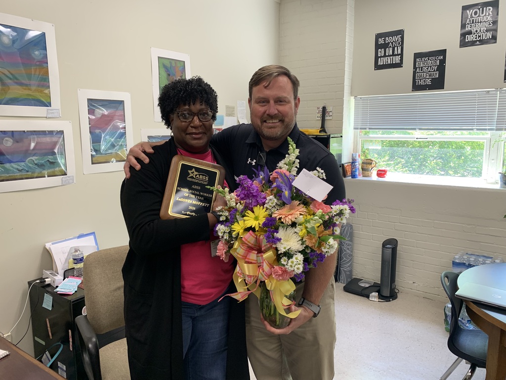 LaSonya standing in her office beside Principal Curry holding a flower bouquet and holding an award plaque