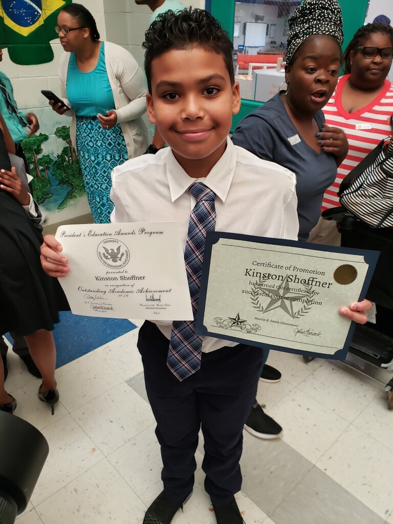 A young boy wearing a white shirt and striped tie holds two award certificates in a school hallway