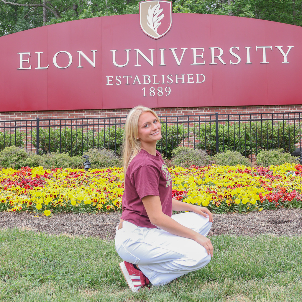 A high school senior squats in front of a floral arrangement and a large Elon University sign