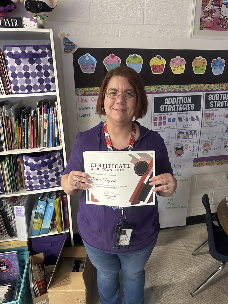 A female teacher with short reddish-brown hair and glasses stands in a classroom holding a certificate of recognition with both hands. She is wearing a purple hoodie, blue jeans, and a Hello Kitty lanyard. Behind her are bookshelves filled with children's books and a bulletin board labeled "Addition Strategies."