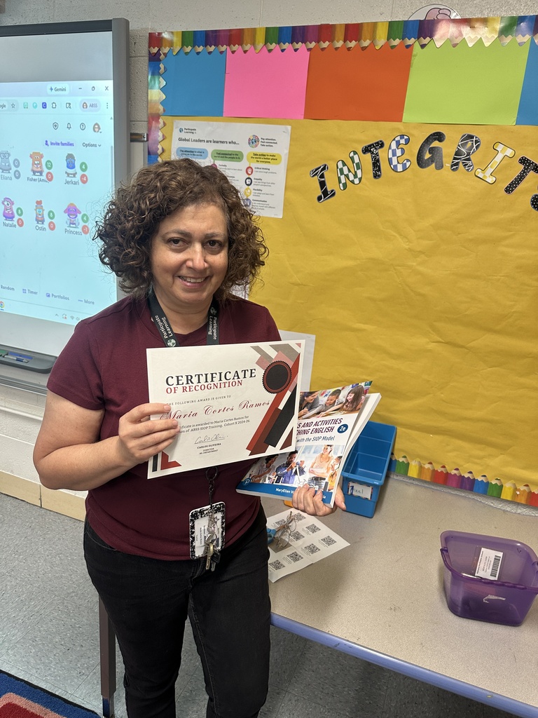 A smiling female teacher with curly brown hair stands in a classroom holding a certificate of recognition and a SIOP instructional book. She is wearing a maroon t-shirt and black jeans. In the background, a yellow bulletin board features the word "INTEGRITY" in colorful decorative letters next to a white interactive whiteboard.