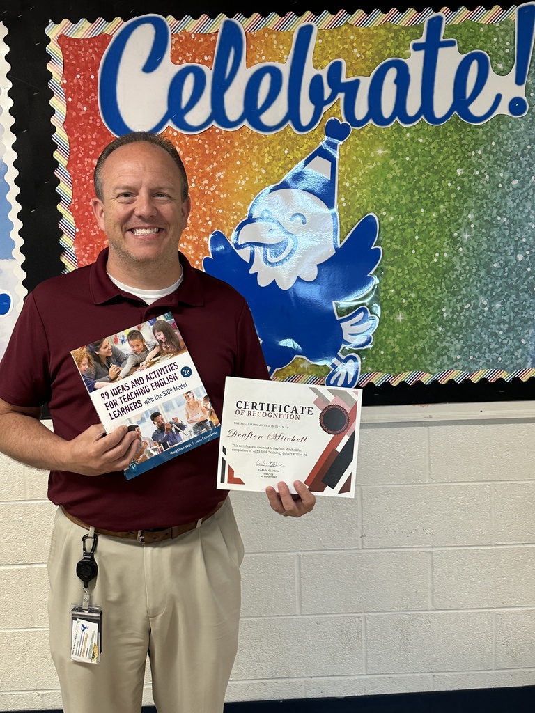 A smiling male teacher with short brown hair stands in front of a colorful school bulletin board. He is wearing a maroon polo shirt and khaki pants, and he is holding a SIOP textbook and a certificate of recognition. The bulletin board behind him displays the word "Celebrate!" above a cartoon illustration of a blue eagle wearing a party hat.