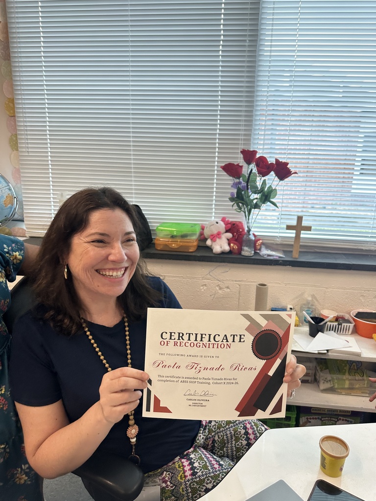 A smiling female teacher with dark hair sits at a desk in a classroom, proudly holding a certificate of recognition. She is wearing a navy blue t-shirt and a beaded necklace with a flower charm. The window behind her has closed blinds, with a small vase of red artificial roses and a wooden cross sitting on the sill.