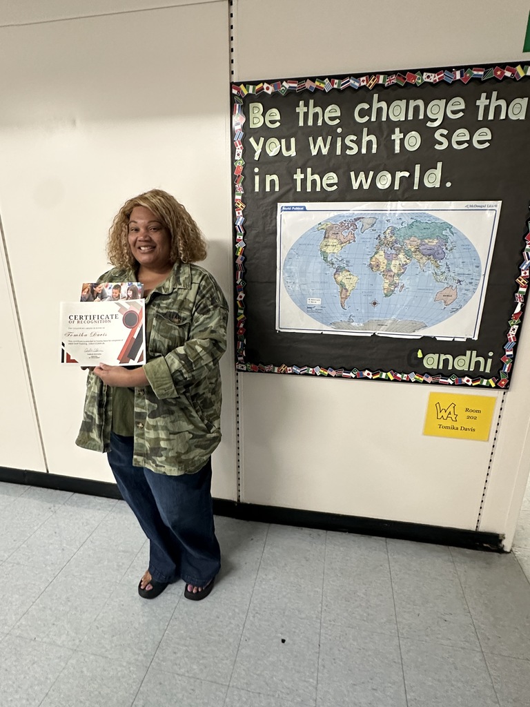 A smiling female teacher with blonde, curly hair stands in a school hallway holding a certificate of recognition. She is wearing a camouflage jacket and wide-leg blue jeans. She stands next to a bulletin board that features a world map and the quote, "Be the change that you wish to see in the world. - Gandhi," alongside a yellow room sign reading "Tomika Davis."