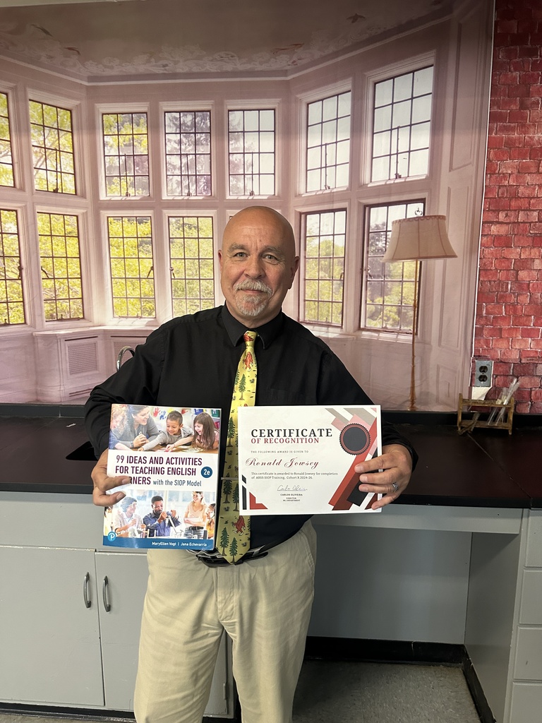 A bald, smiling male teacher wearing a black button-down shirt and a bright yellow tie patterned with green trees and red designs stands in a classroom. He holds up a certificate of recognition in his left hand and a textbook titled "99 Ideas and Activities for Teaching English Learners with the SIOP Model" in his right hand. The background features a large wall mural depicting a white room with large bay windows looking out onto trees.