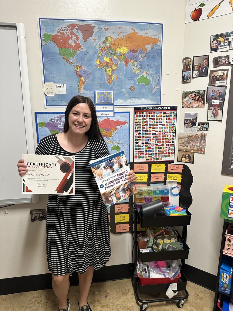 A smiling female teacher with long dark hair stands in a classroom holding a certificate of recognition and a SIOP instructional textbook. She is wearing a black-and-white striped dress. Behind her is a white wall featuring a large map of the world, a "Flags of the World" poster, and a collage of family photographs.