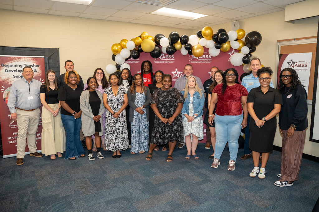 Large group photo of teachers in front of a ABSS maroon backdrop with black, gold, and white balloons