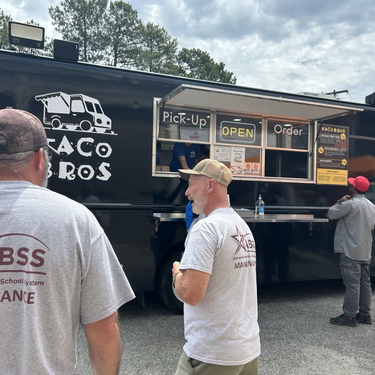 Maintenance workers wearing gray tshirts stand in front of a black Taco Bros food truck waiting for their food