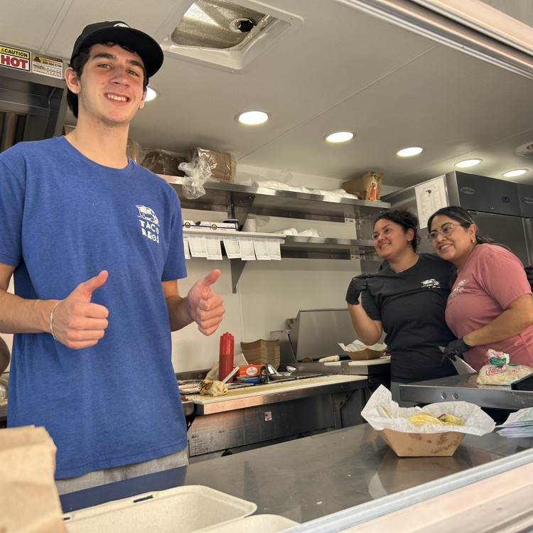 Three taco truck employees smile towards the camera toward a very busy lunch service