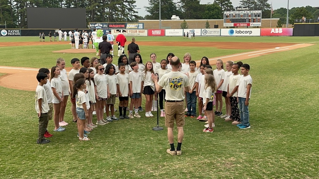 A large group of elementary students in tan shirts with music teacher directing them, standing on a baseball field singing the National Anthem