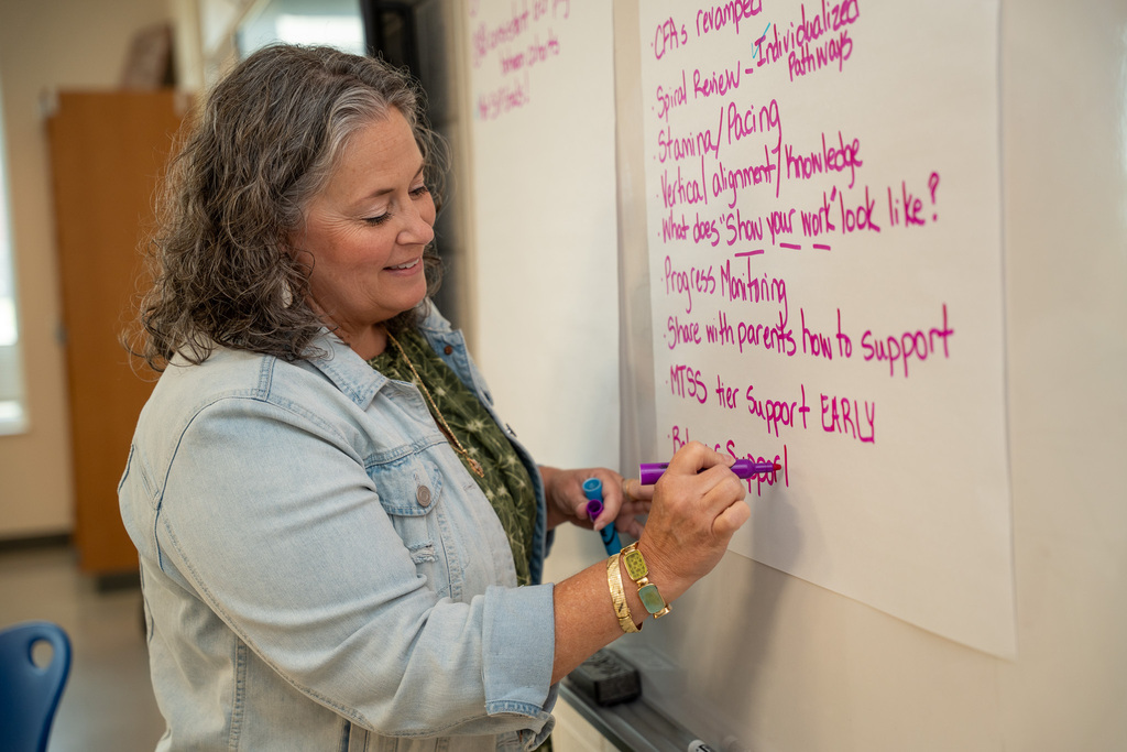 A medium shot of a smiling woman with wavy gray hair wearing a denim jacket, writing bullet points with a purple marker on a large sheet of chart paper attached to a whiteboard.