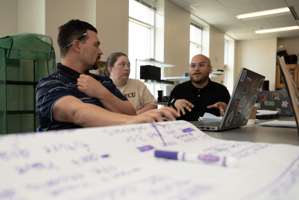 A low-angle shot focusing on three educators sitting at a table engaged in discussion. A man in a striped shirt listens as another man with glasses and a black shirt gestures while speaking next to an open laptop. In the foreground, a large sheet of paper with handwritten notes and a purple marker is softly blurred.