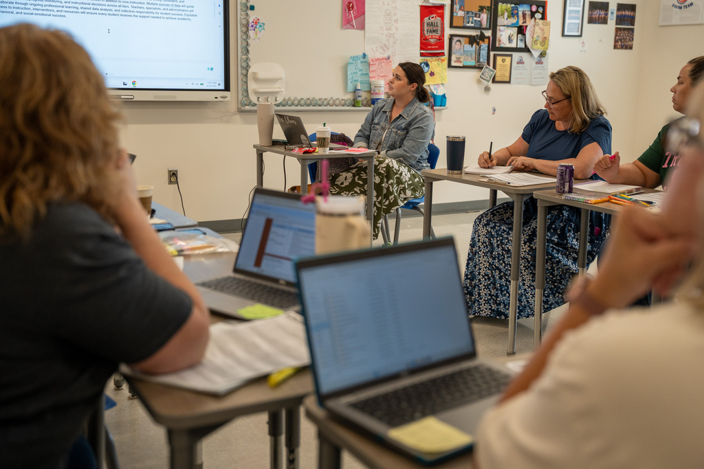 A classroom setting showing educators collaborating during a meeting. In the foreground, the backs of two participants are visible next to open laptops. In the background, three women are seated at desks; one in a denim jacket looks toward a large white interactive screen, while another writes in a notebook.