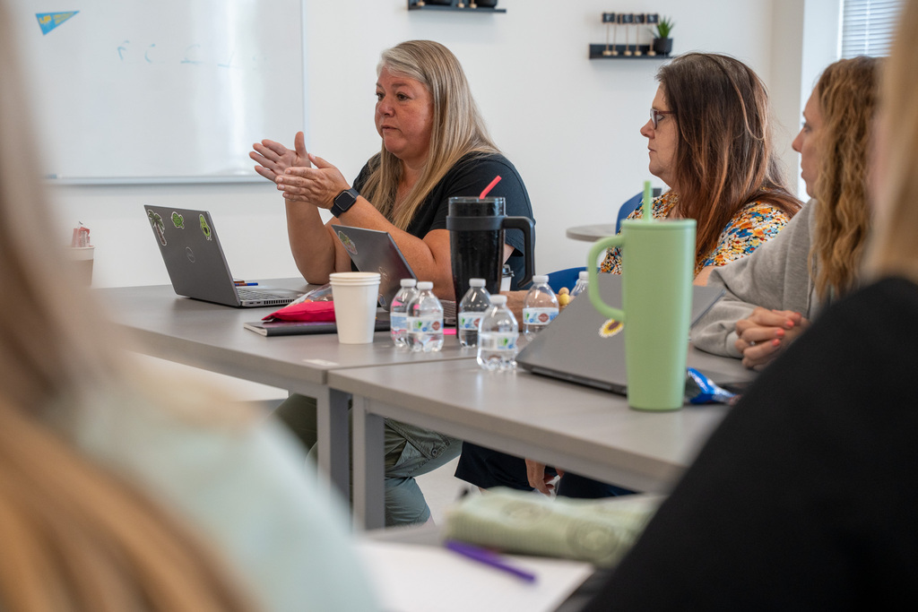 A close-up, side-angle shot of a woman with long blonde hair speaking and gesturing with her hands while seated at a conference table. Other participants sit beside her with laptops and water bottles, listening intently during a group discussion.