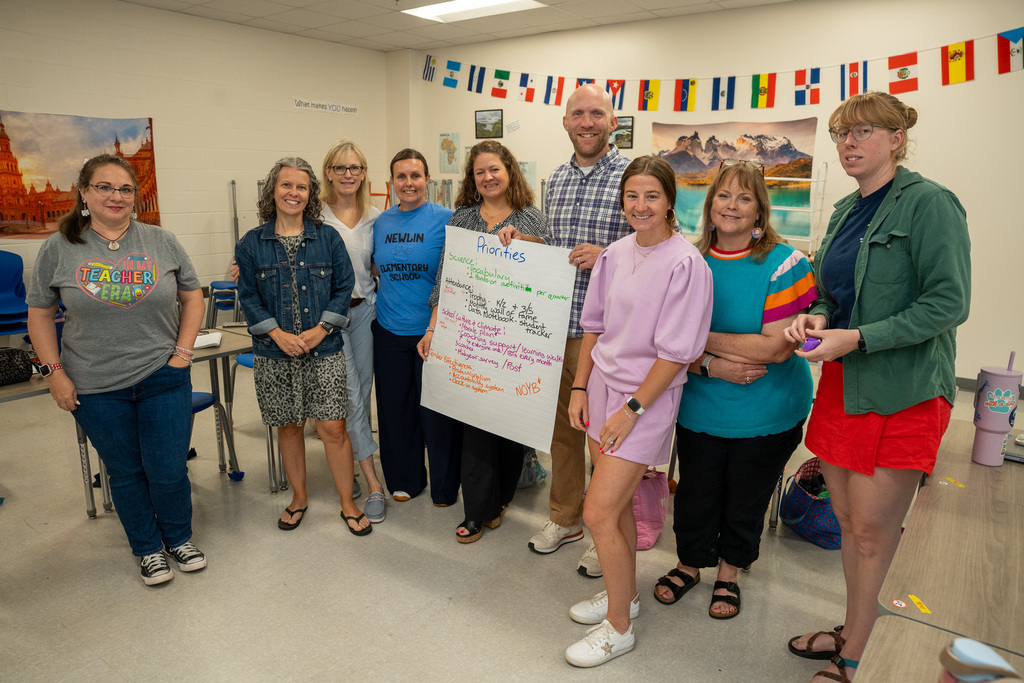 A group of nine educators standing together in a classroom, smiling for a group photo while holding up a large sheet of chart paper titled "Priorities" with handwritten notes. A string of international flags hangs on the wall behind them.