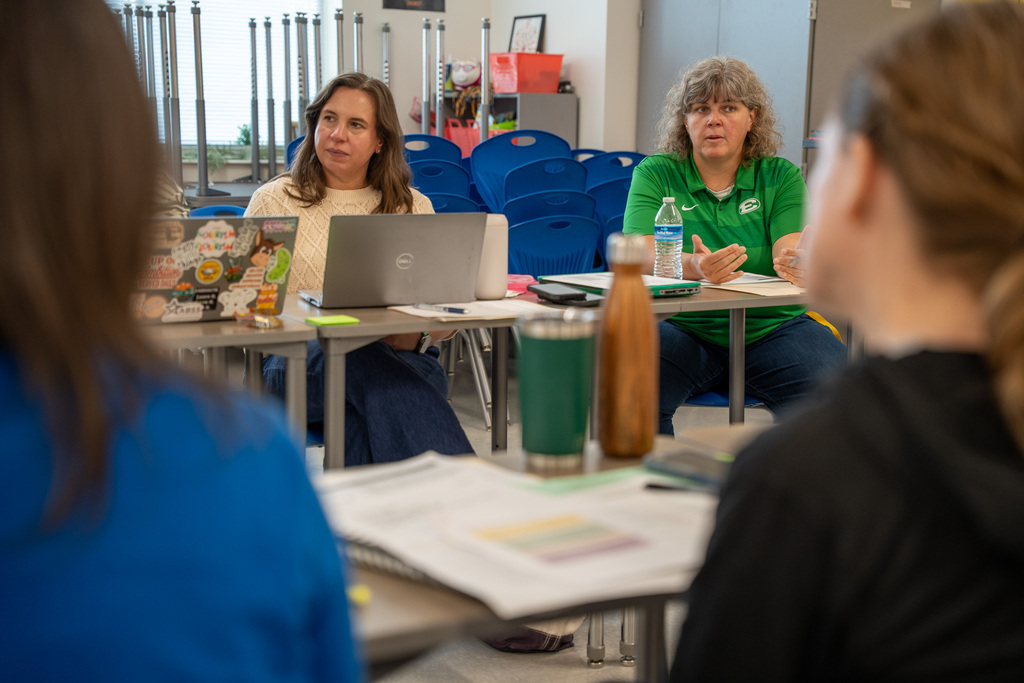 A classroom discussion view from behind a participant. Two women are seated at desks in the middle ground facing forward; one woman in a cream sweater sits next to a laptop, while another woman in a green shirt gestures with her hands as she speaks to the room.