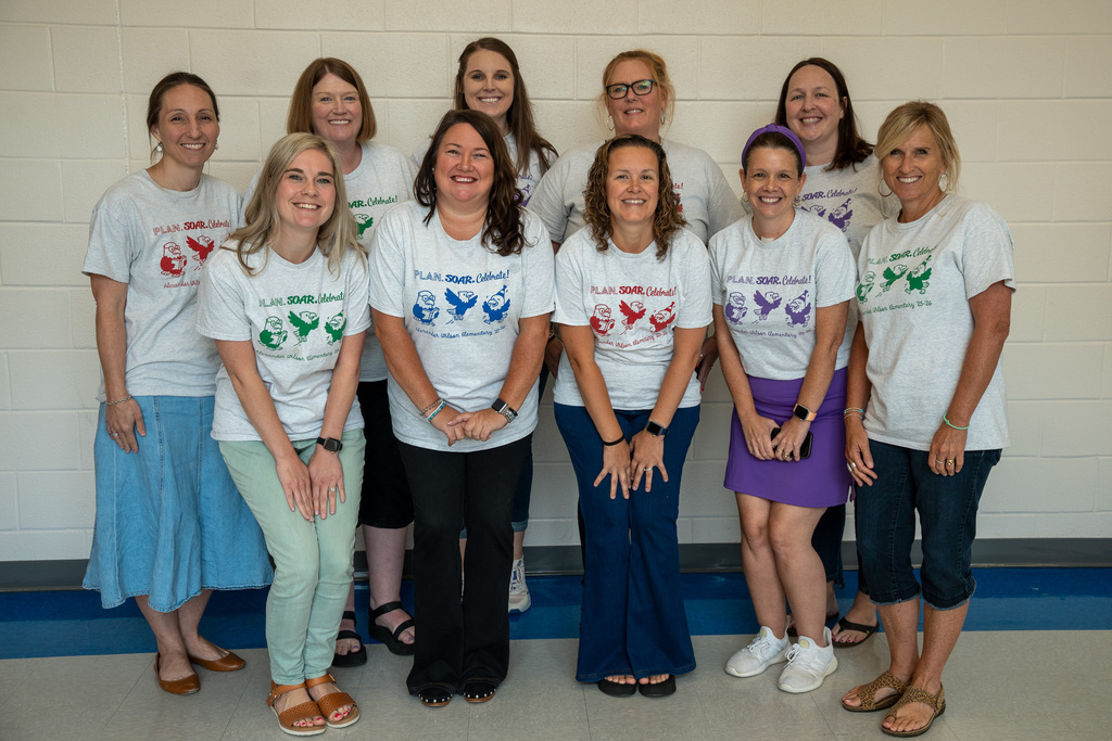 A group photo of nine smiling women standing against a white brick wall indoors. Most of them are wearing matching light-gray t-shirts that feature a graphic of cartoon birds and the text "PLAN. SOAR. Celebrate!"