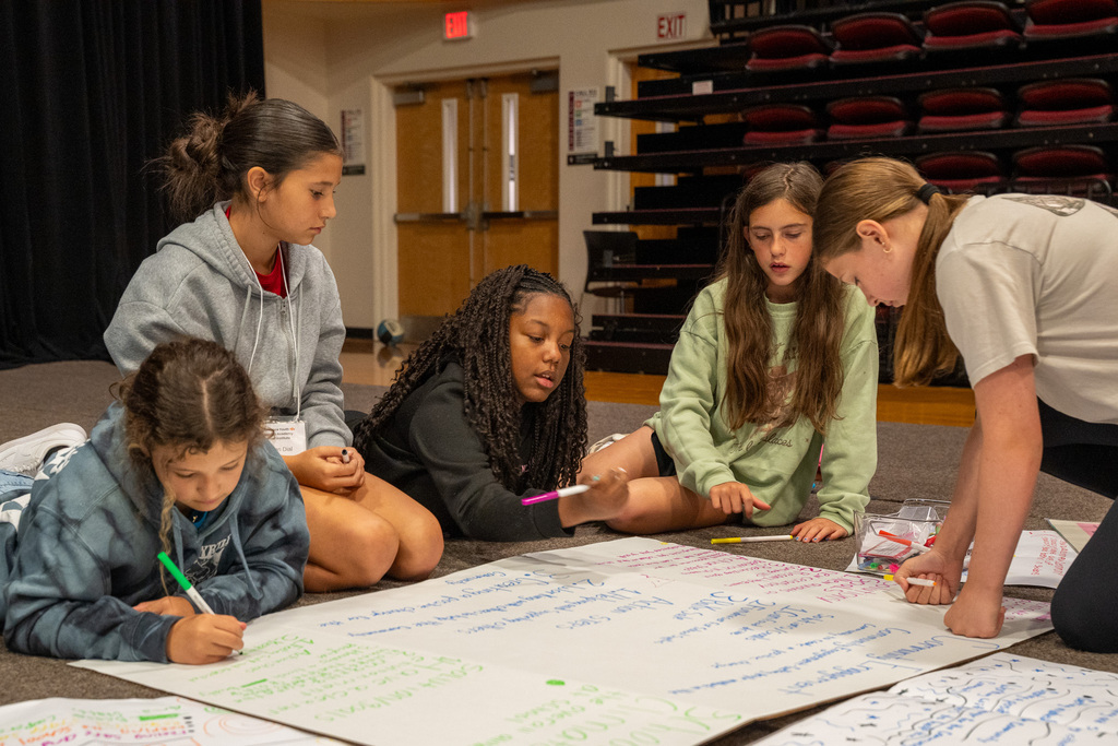 Five middle school girls gather around a large poster paper for a group project