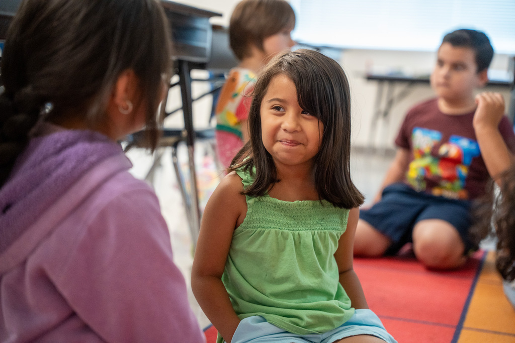 An elementary student in a green shirt smirks at another student during a partner talk moment on a classroom carpet