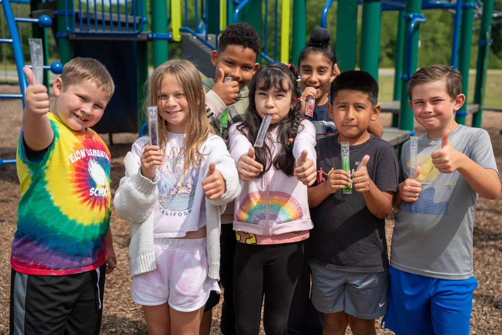 A group of elementary students giving thumbs up on a playground. Some are eating an frozen popsicle treat.