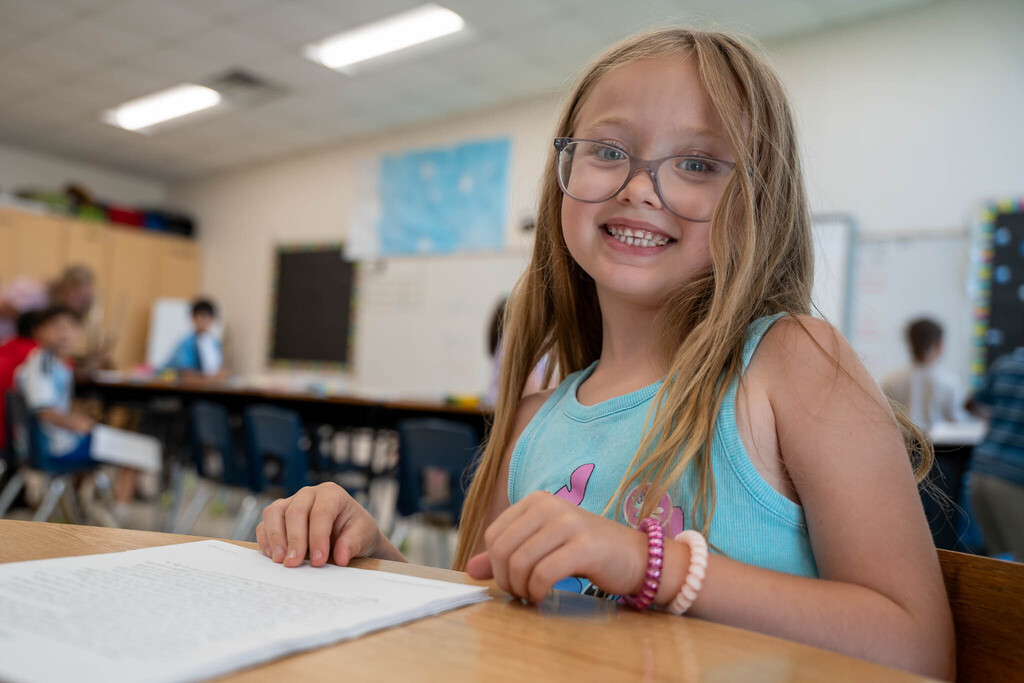 A girl in a blue tank top and pink bracelets looks up and smiles with a reading passage in front of her