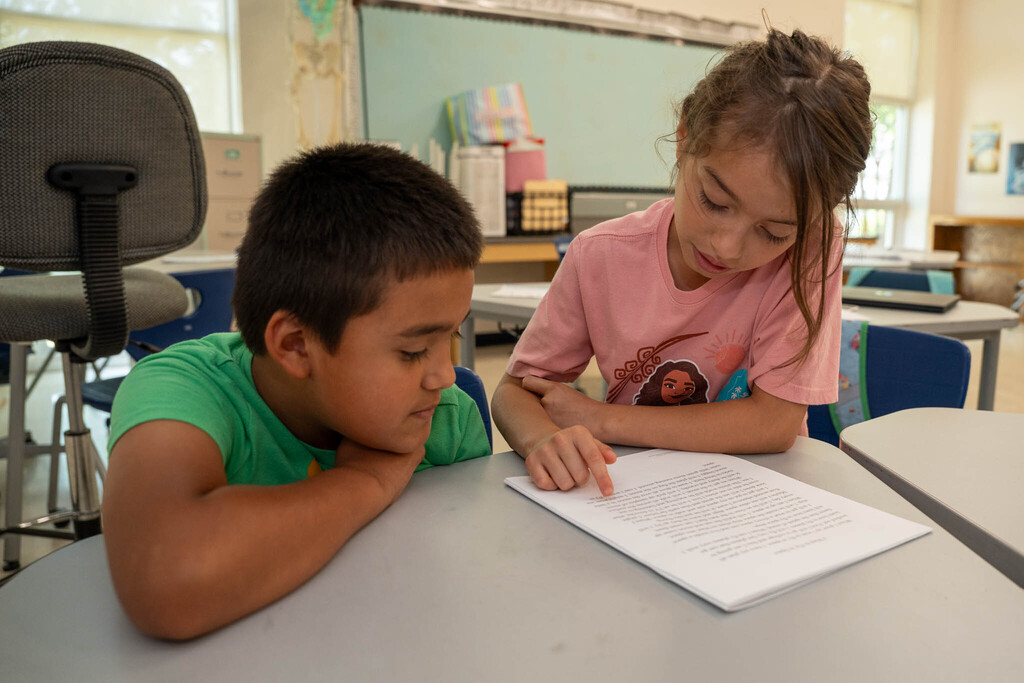 A girl in a pink Moana shirt points to a word as she reads with another student in a green t-shirt looking closely beside her