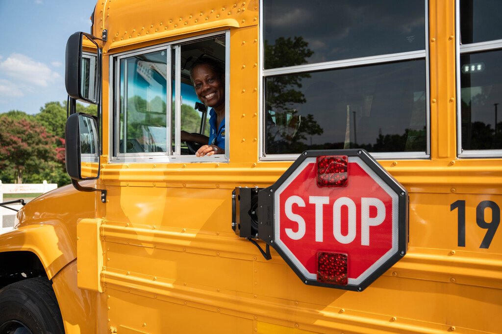 A bus driver in a blue polo leans out the window of a brand new yellow school bus and smiles