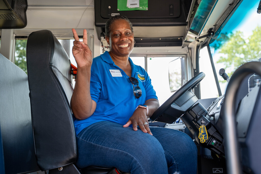 A bus driver in a blue polo shirt gives a peace sign and smiles behind the wheel of a brand new school bus