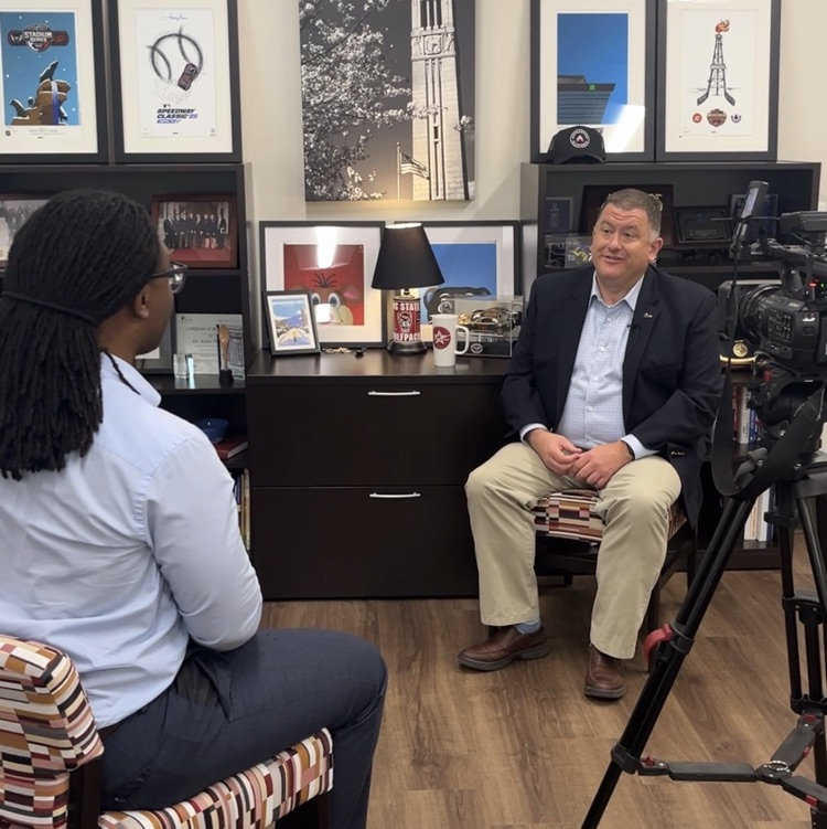 A news anchor sits in front of the superintendent in his office to discuss back to school updates. A camera to the right points at the superintendent.