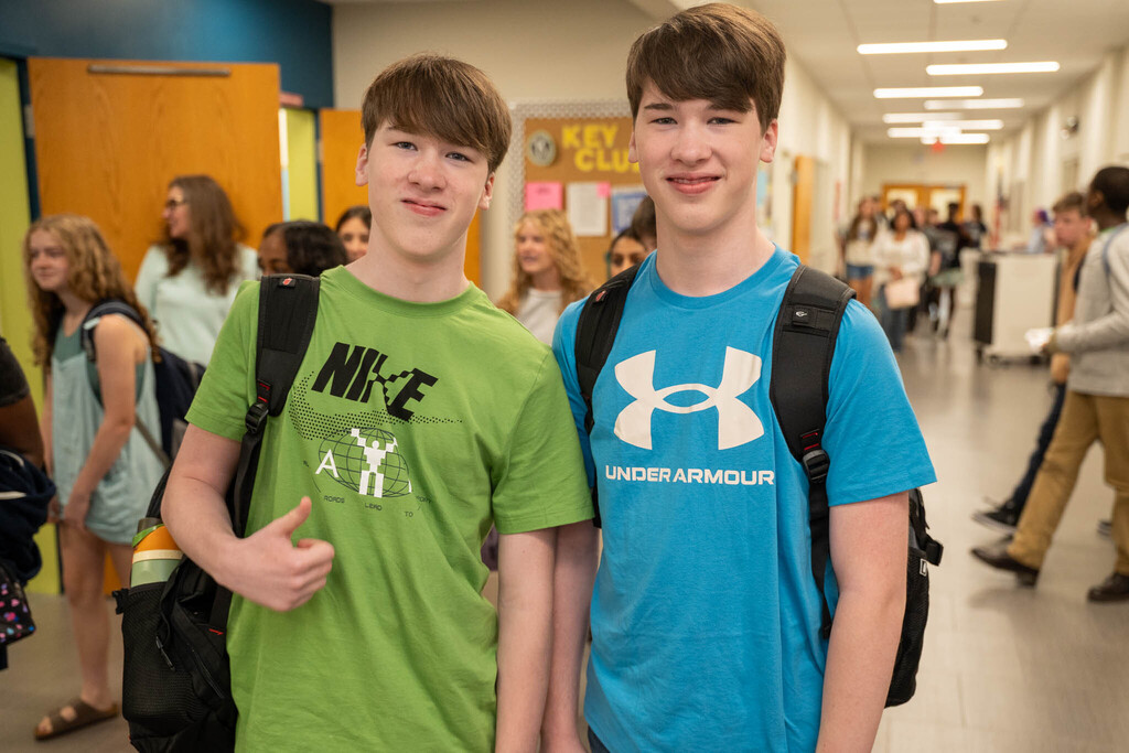 Two young male students with similar dark brown haircuts stand in a school hallway. One wears a green t-shirt with a Nike logo, giving a thumbs up. The other wears a blue t-shirt with an Under Armour logo. They both wear backpacks. Other students are visible in the background.
