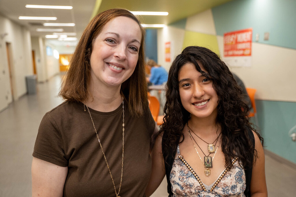 A photo of a female teacher in a brown t-shirt and a student with long curly dark hair wearing a patterned dress and multiple necklaces. Both are smiling warmly as they stand together in a brightly lit school hallway with classroom doors and posters on the walls.