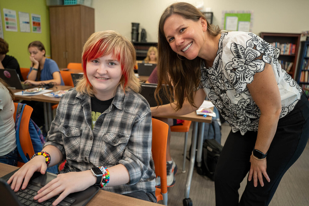 A smiling female teacher in a white top with black lace patterns leans over a student who is sitting at a desk and looking up. The student has curly hair with red highlights and is wearing a plaid shirt. They are surrounded by other students working on laptops in a busy, colorful classroom.