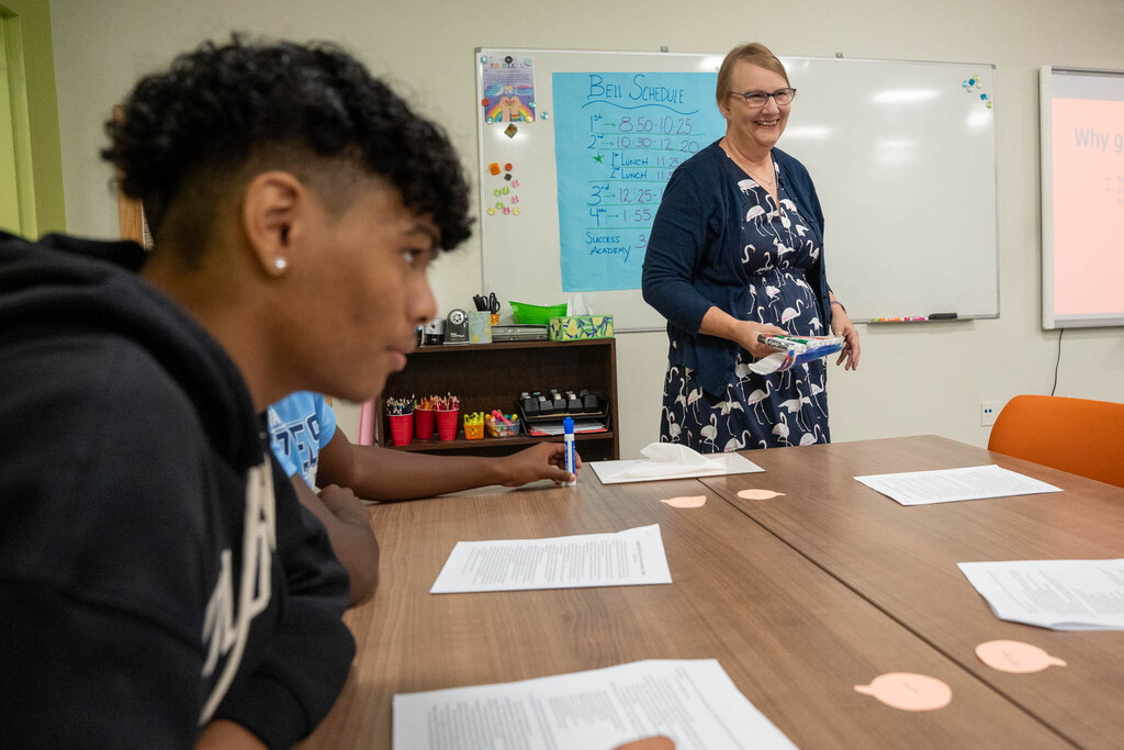A classroom shot focused on a student with a close-cropped haircut, viewed mostly from behind and to the side, looking towards a smiling female teacher in a navy dress standing at a desk holding a container of markers. The background shows a white board and posters, including a handwritten bell schedule.