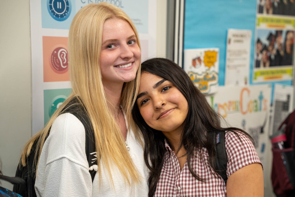 A photo of two smiling high school female students posing closely in a hallway. One student has long blonde hair and is wearing a white sweatshirt. The other student has long dark hair, wearing a plaid shirt, and is resting her head on the blonde student's shoulder. Colorful posters line the wall behind them.