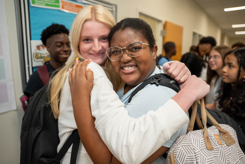Two diverse young women, both wearing backpacks, share a cheerful hug in a crowded school hallway. The student on the left has blonde hair and a white sweatshirt. The student on the right, wearing glasses, a light-colored top, and holding a patterned tote bag, has a wide-eyed, joyful expression. Other students fill the background.