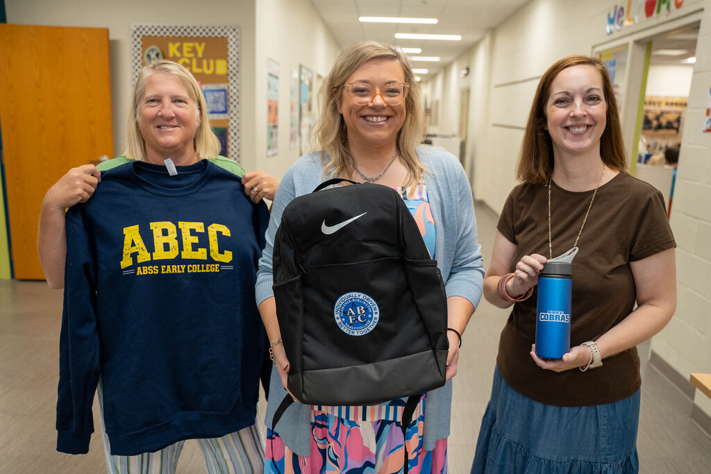 Three smiling women stand in a school hallway holding up ABEC spirit wear. From left to right: one woman holds a navy blue sweatshirt with yellow text reading "ABEC ABSS EARLY COLLEGE"; the middle woman holds a black Nike backpack; and the right woman holds a blue water bottle. In the background, there's a "KEY CLUB" bulletin board.