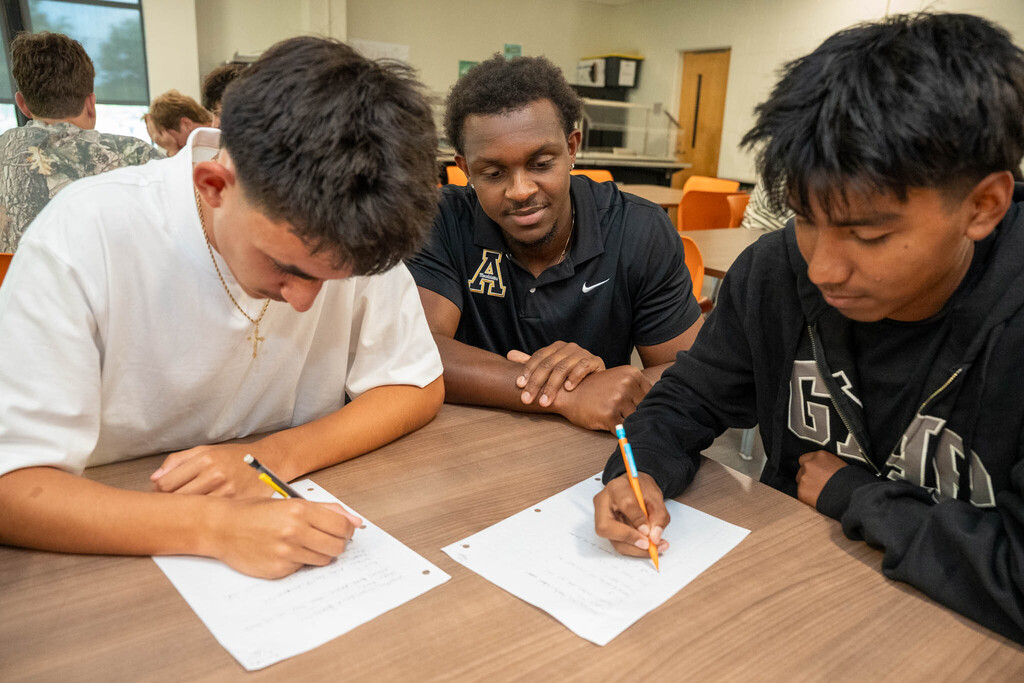 Three people sitting at a wooden desk in what appears to be a classroom. Two young men are leaning forward and writing with pencils on white paper, while an older Black man, wearing a polo shirt with an 'A' logo, watches them with a gentle expression.