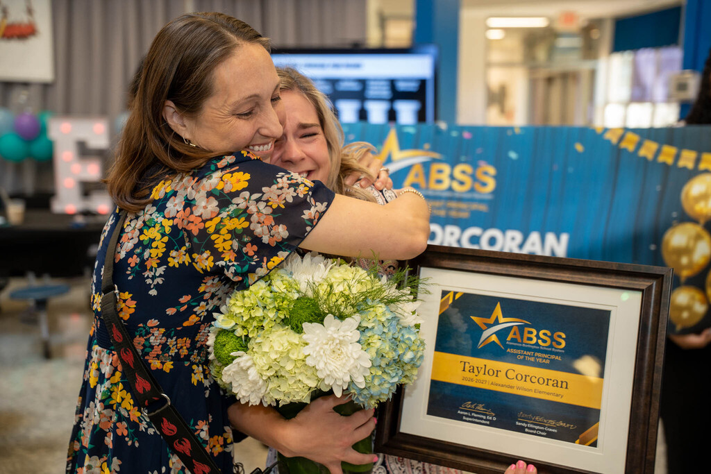 A colleague embraces an emotional Taylor Corcoran, who holds a floral arrangement and framed award certificate.