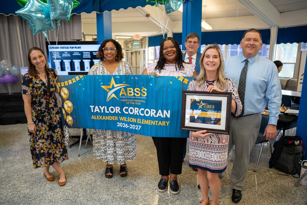 Taylor Corcoran stands in the center of school administrators and district officials who are holding a custom recognition banner and framed certificate.