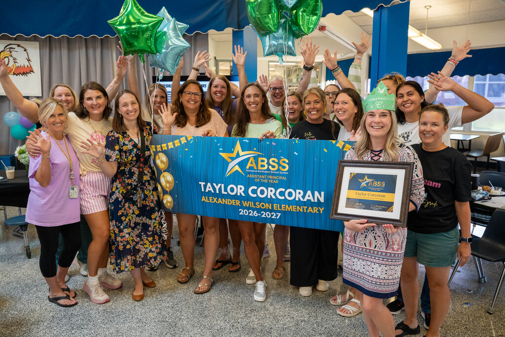 A large group of elementary school staff members poses together, waving and cheering behind a recognition banner with Taylor Corcoran in the foreground holding her award.