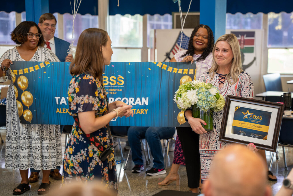 Taylor Corcoran holds her award certificate and flowers while listening to a speaker in front of a blue celebration banner reading "ABSS Assistant Principal of the Year."