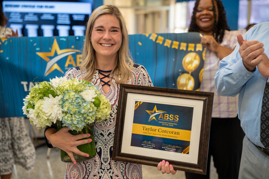 Taylor Corcoran smiles while holding a framed award certificate for "ABSS Assistant Principal of the Year 2026-2027" and a bouquet of flowers.