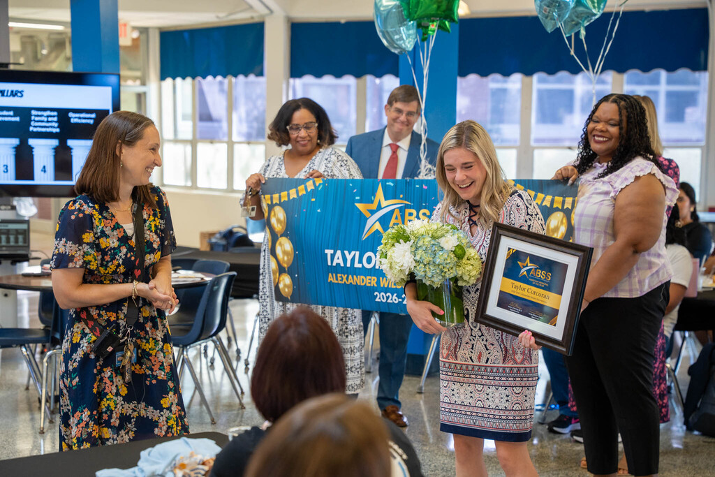 Taylor Corcoran laughs while holding her framed award certificate and flowers next to colleagues holding a congratulatory banner.