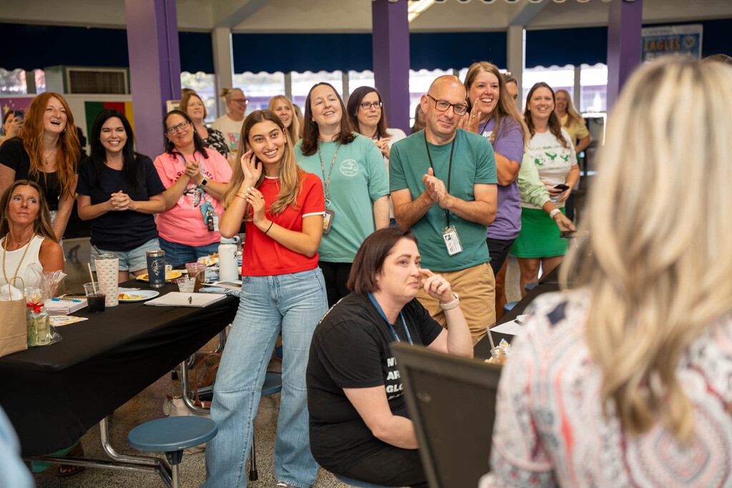 A crowd of elementary school staff members smiles and applauds during a recognition presentation.