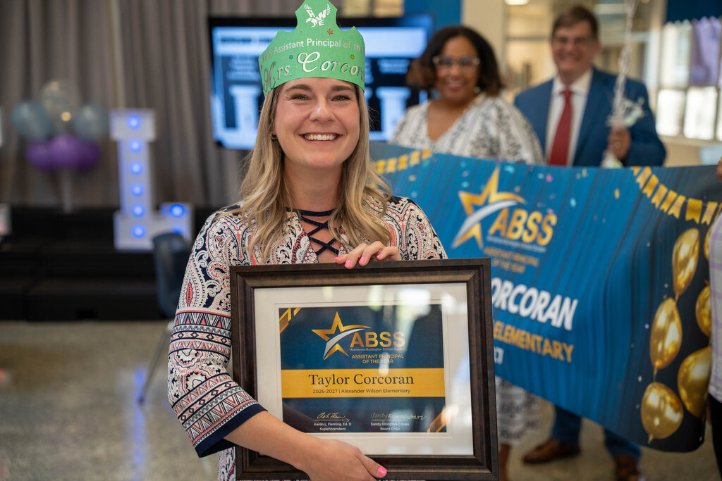 Taylor Corcoran wears a green paper crown that reads "Mrs. Corcoran" while holding her framed Assistant Principal of the Year certificate.