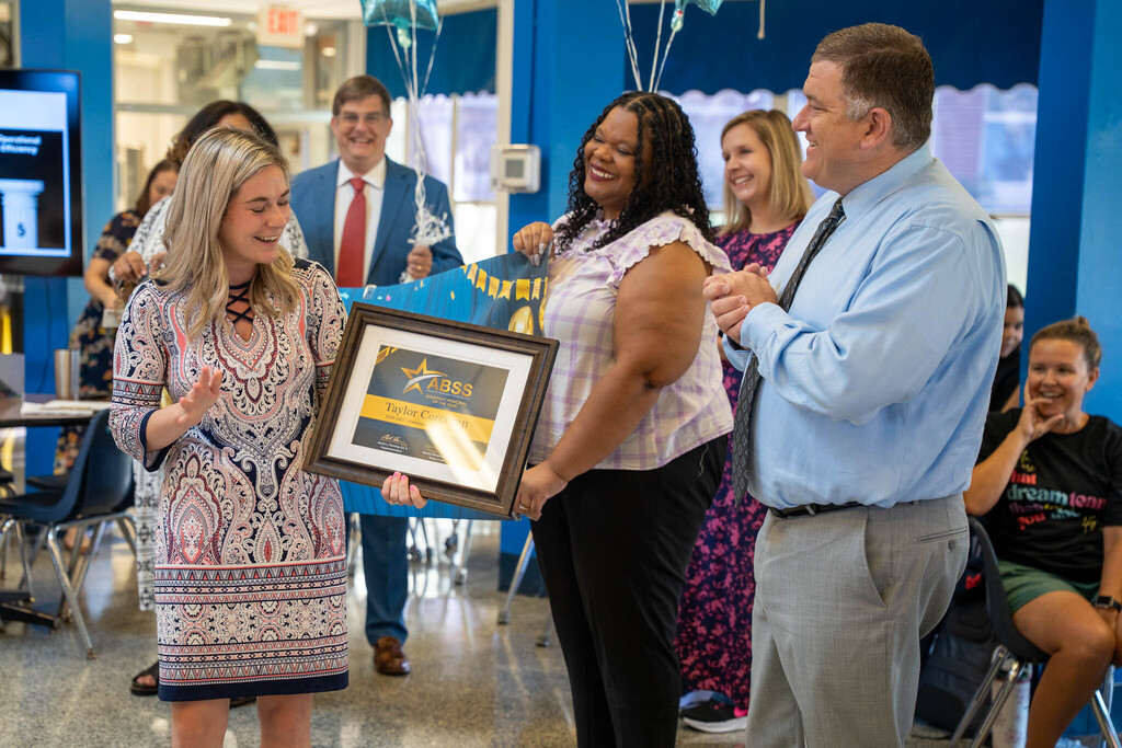 Taylor Corcoran reacts joyfully as colleagues and school administrators present her with an award certificate and congratulatory banner.