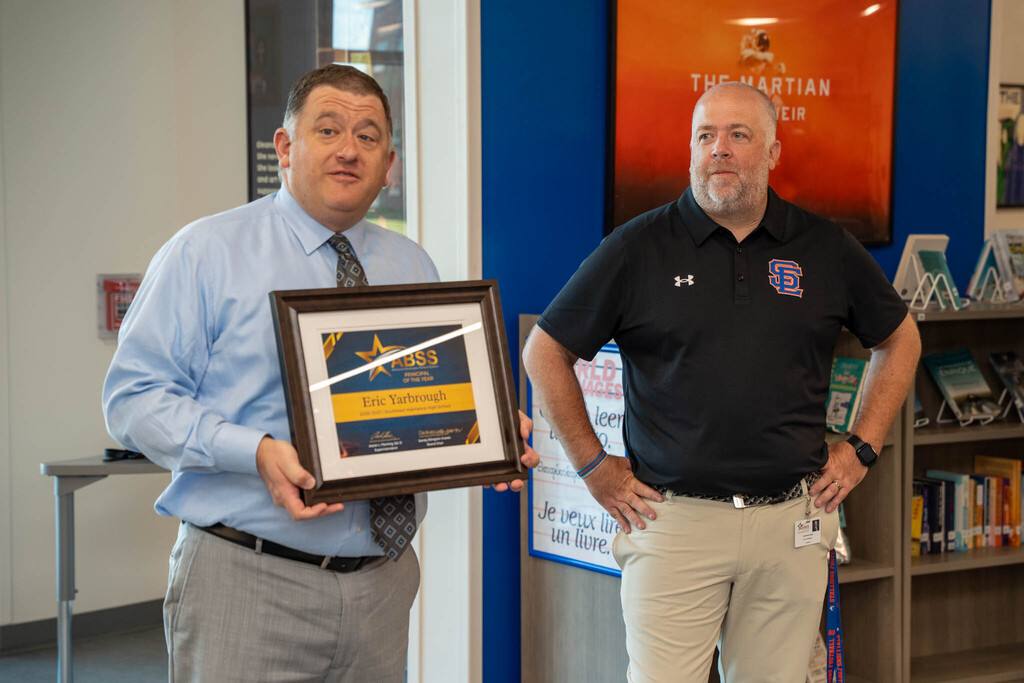A district administrator holds a framed certificate for "ABSS Principal of the Year" next to Principal Eric Yarbrough, who stands with hands on his hips.